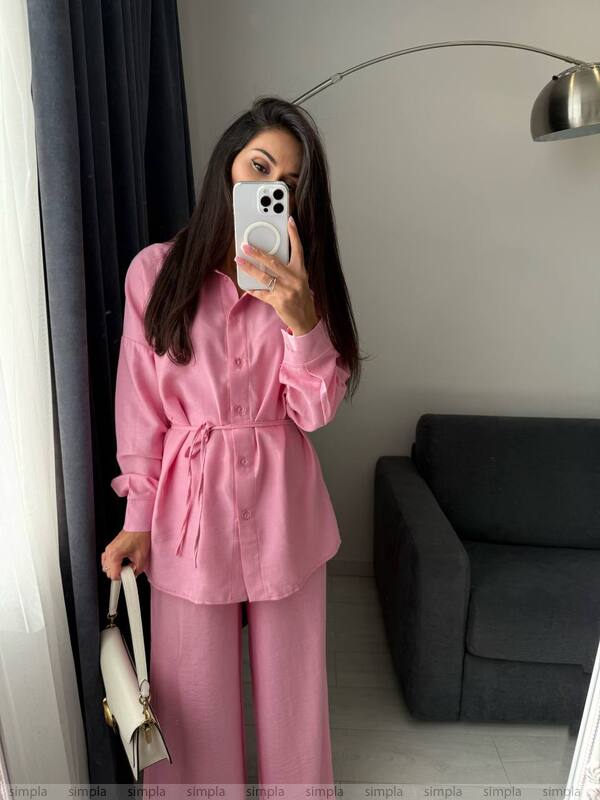 Person wearing a pink outfit taking a mirror selfie in a room with a gray sofa and lamp.