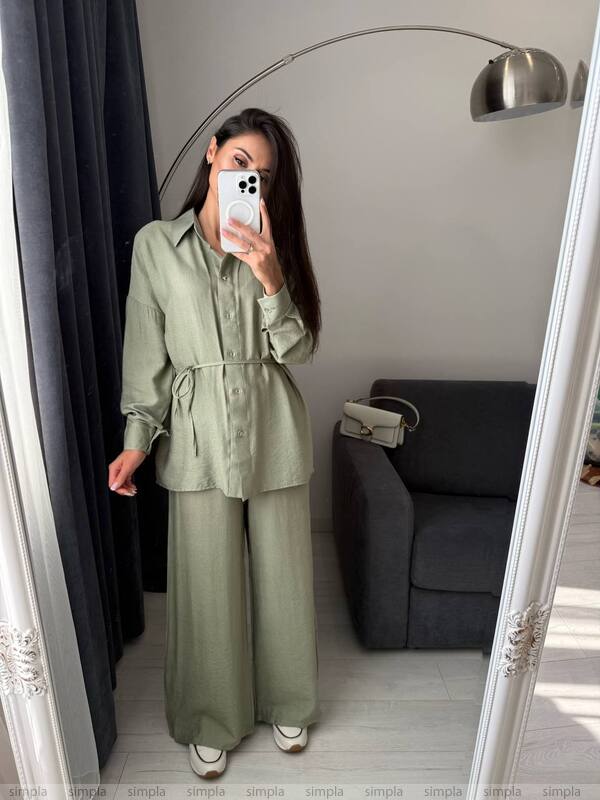Person wearing a green outfit taking a mirror selfie in a room with a gray sofa and lamp.