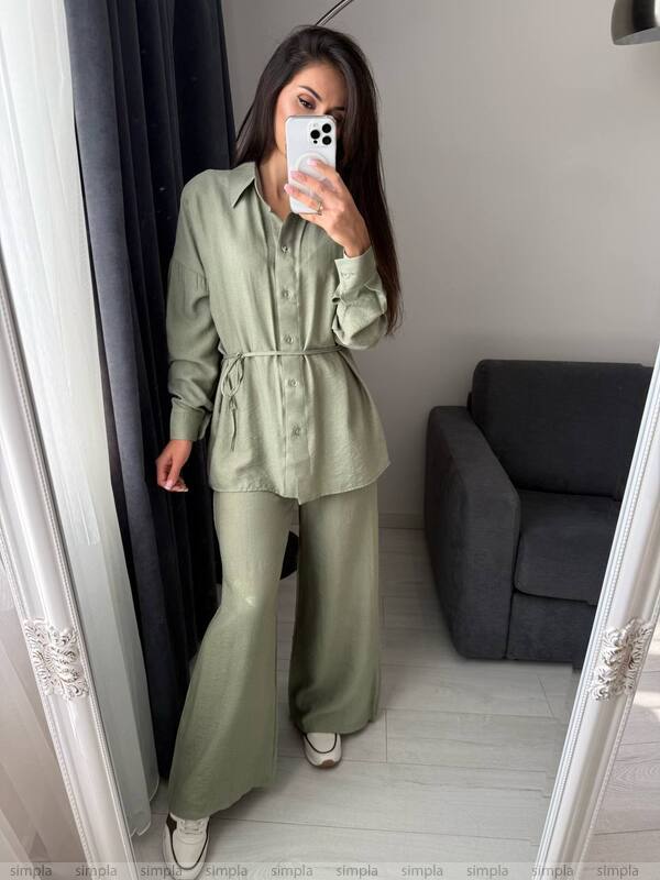 Woman taking a mirror selfie wearing a green outfit in a room with a gray sofa.