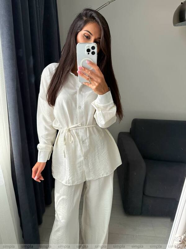 Woman taking a mirror selfie wearing a white outfit in a room with a gray sofa and dark curtains.