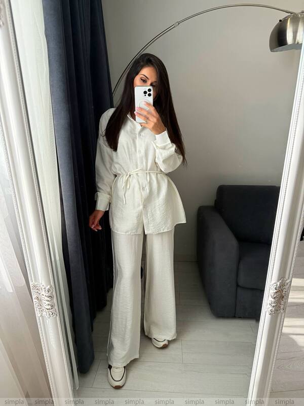 Person wearing a white outfit taking a mirror selfie in a room with a sofa and lamp.