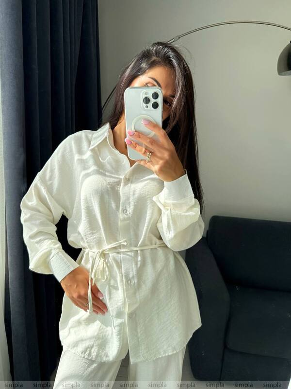 Person wearing a white outfit taking a mirror selfie in a room with dark curtains and a gray sofa.