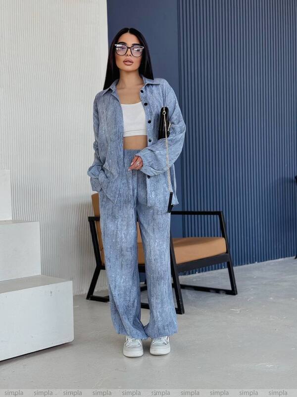 Woman wearing a blue denim outfit standing in an indoor setting with a chair and wall in the background.