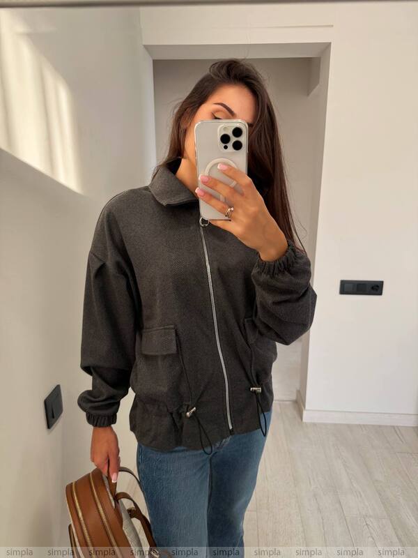Woman taking a mirror selfie wearing a dark jacket and blue jeans in a room.