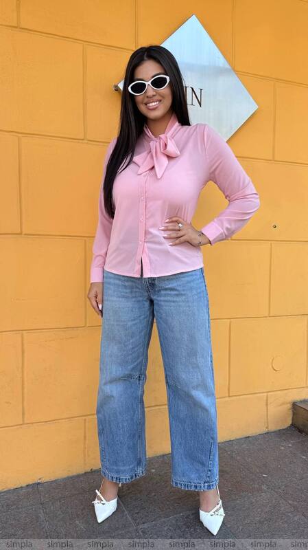 Woman wearing a pink blouse with a bow tie and blue jeans against a yellow wall.
