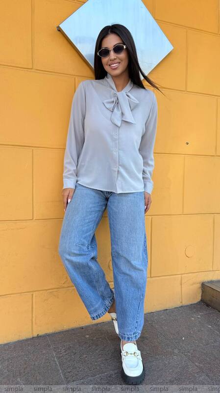 Woman wearing a light gray blouse with a bow tie and blue jeans against a yellow wall.