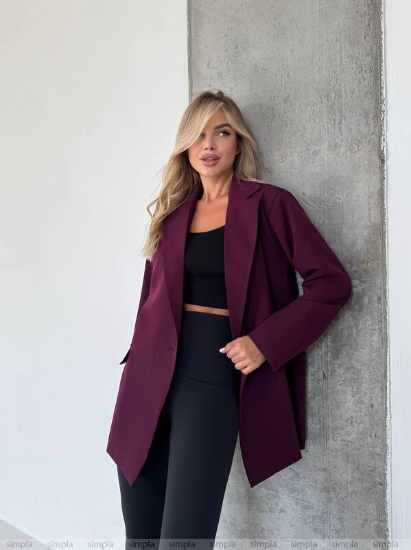 Woman wearing a purple coat and black outfit leaning against a gray wall.