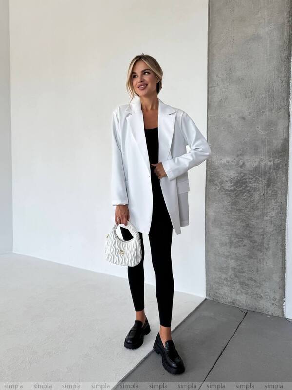 Woman in a white coat and black pants holding a white handbag against a plain wall.