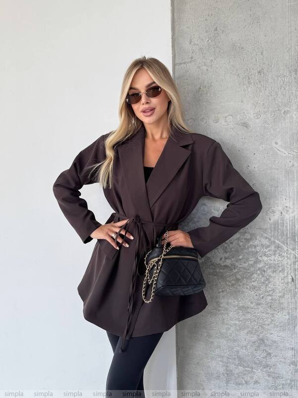 Woman wearing a brown coat and sunglasses, standing against a light gray wall.