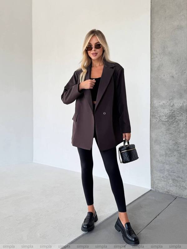 Woman in a brown blazer and black outfit holding a small black handbag against a plain background