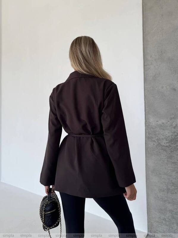 Person wearing a dark brown coat standing against a white wall.