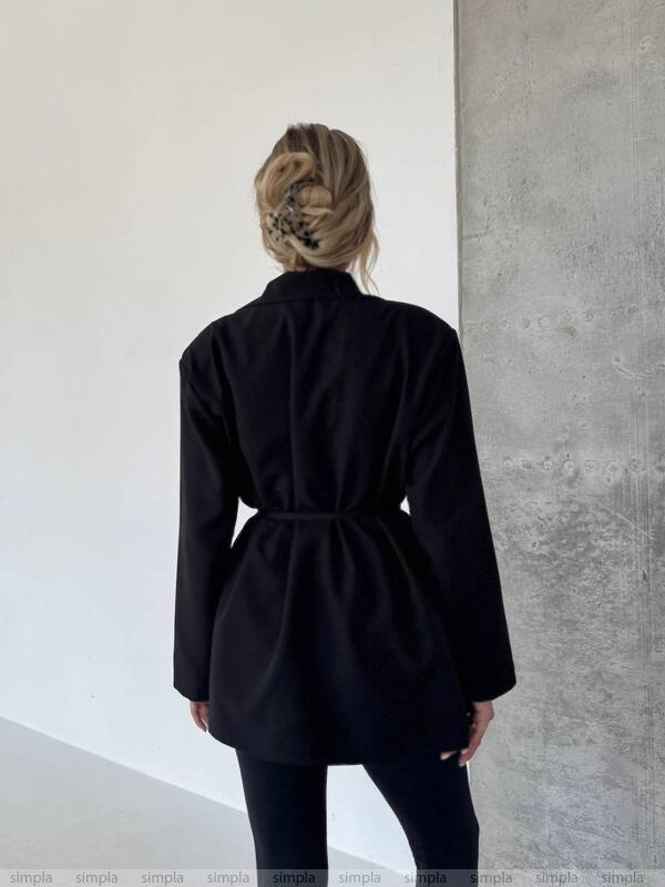 Person wearing a black coat standing against a white and gray wall.