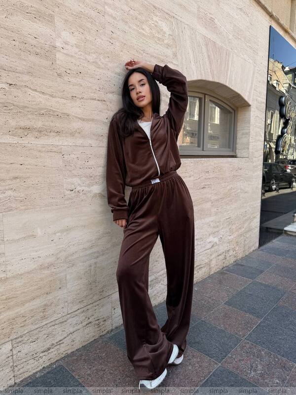 Woman in a brown tracksuit leaning against a stone wall.