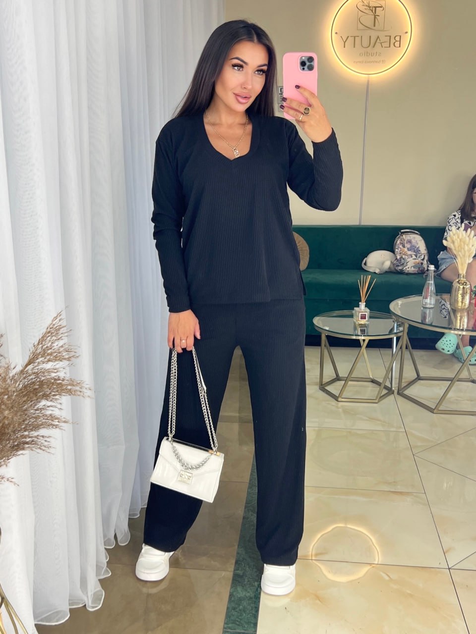 Woman taking a mirror selfie wearing a black outfit with a white handbag in a living room.