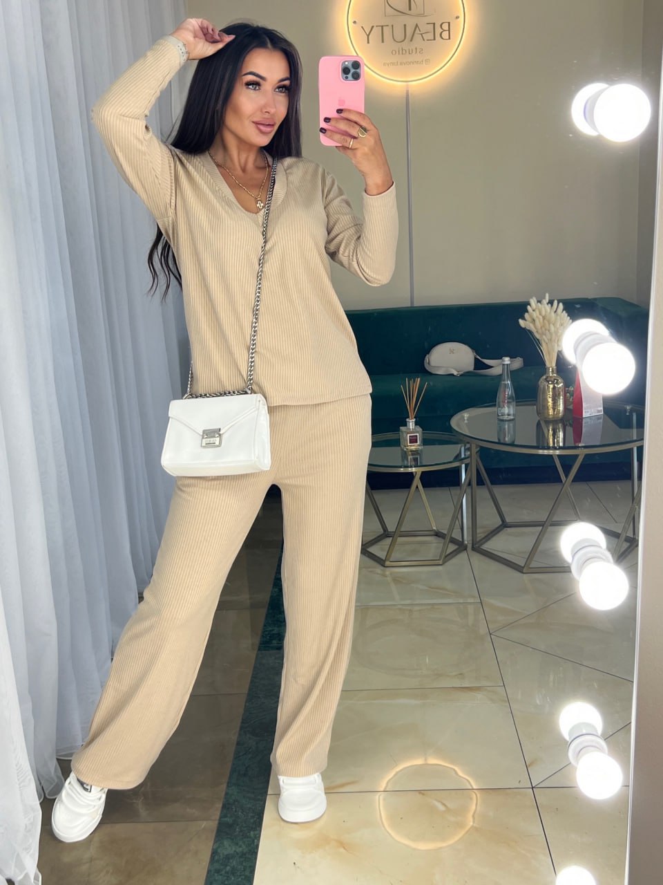 Woman taking a mirror selfie wearing a beige outfit with a white handbag in a well-lit room.