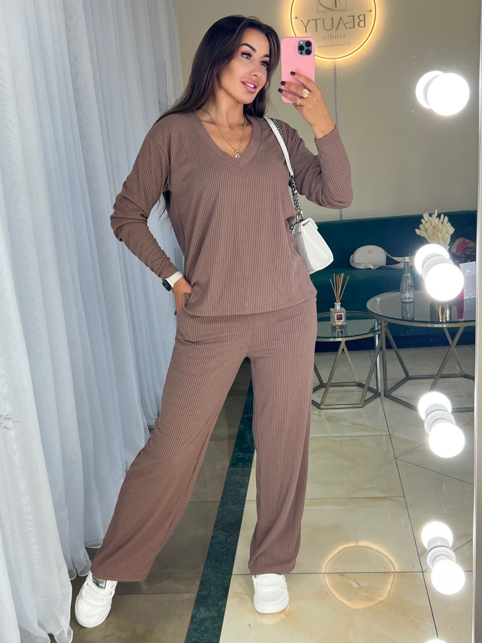 Woman taking a mirror selfie wearing a brown outfit in a room with a blue rug and white curtains.
