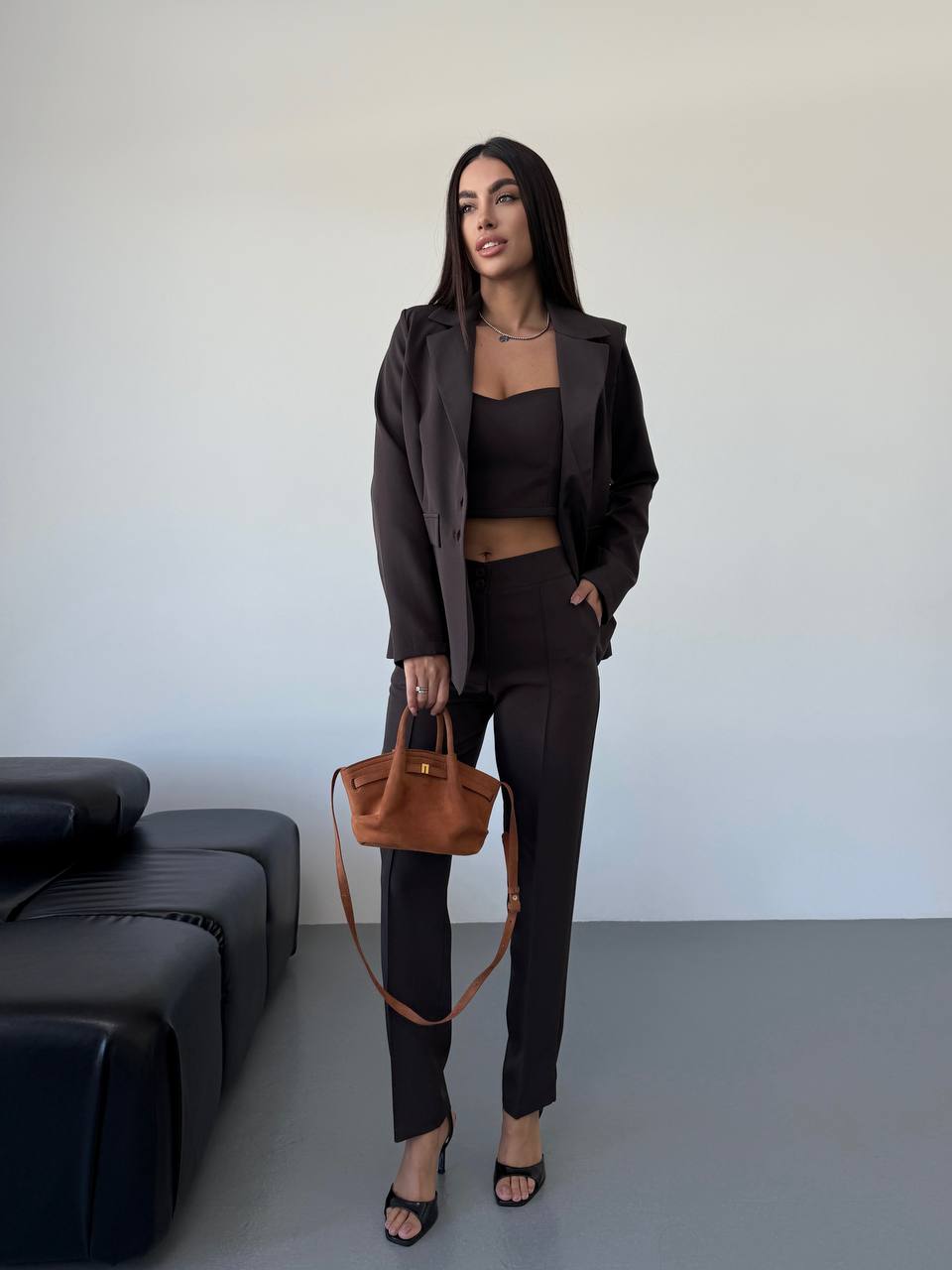 Woman in a stylish outfit with a brown jacket and pants, holding a brown handbag against a plain background.