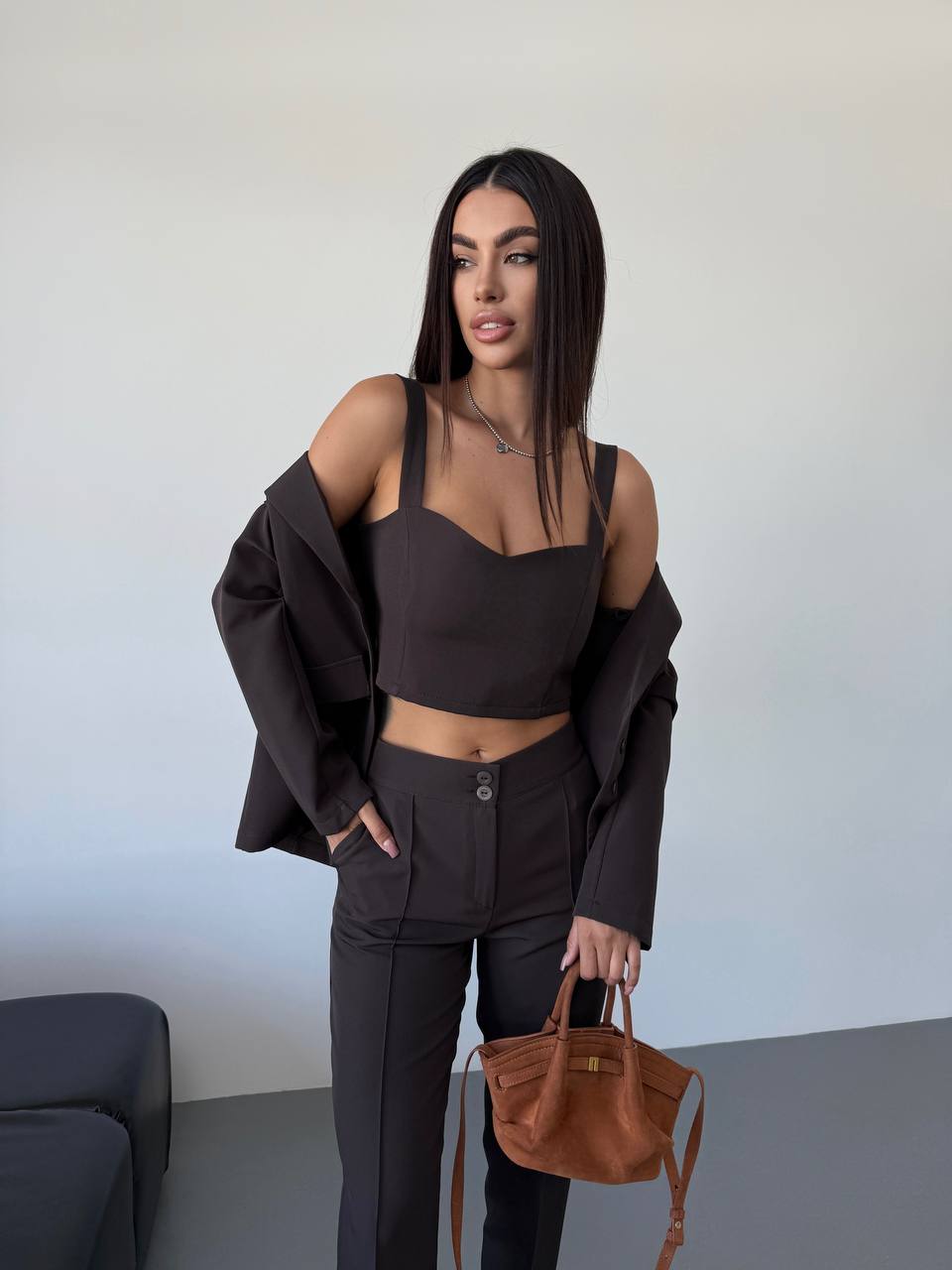 Woman in a dark outfit holding a brown handbag against a plain background