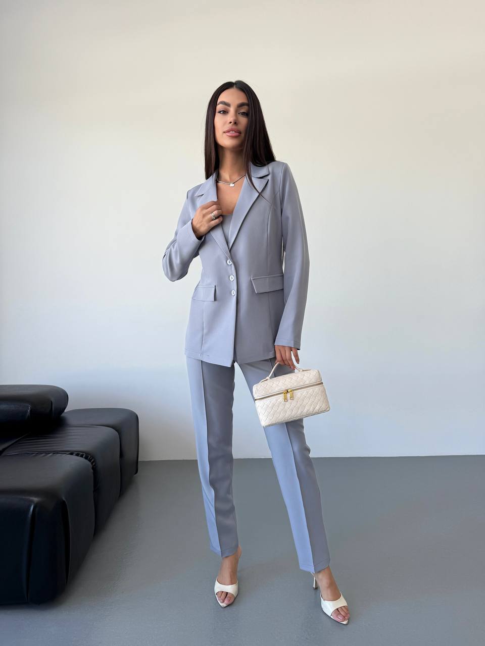 Woman in a gray suit holding a white handbag in an indoor setting.