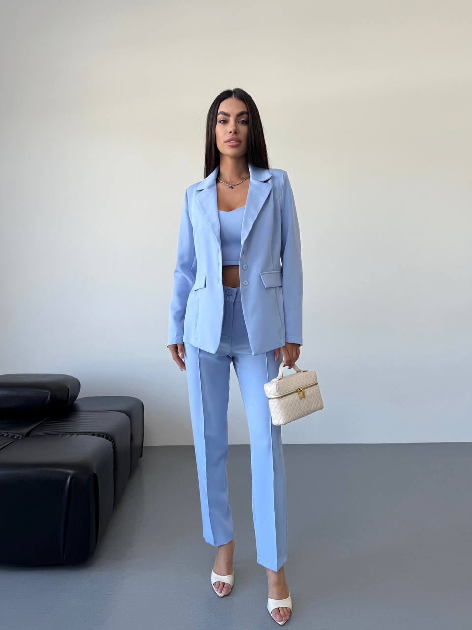 Woman in a light blue suit holding a beige handbag in a minimalistic room.