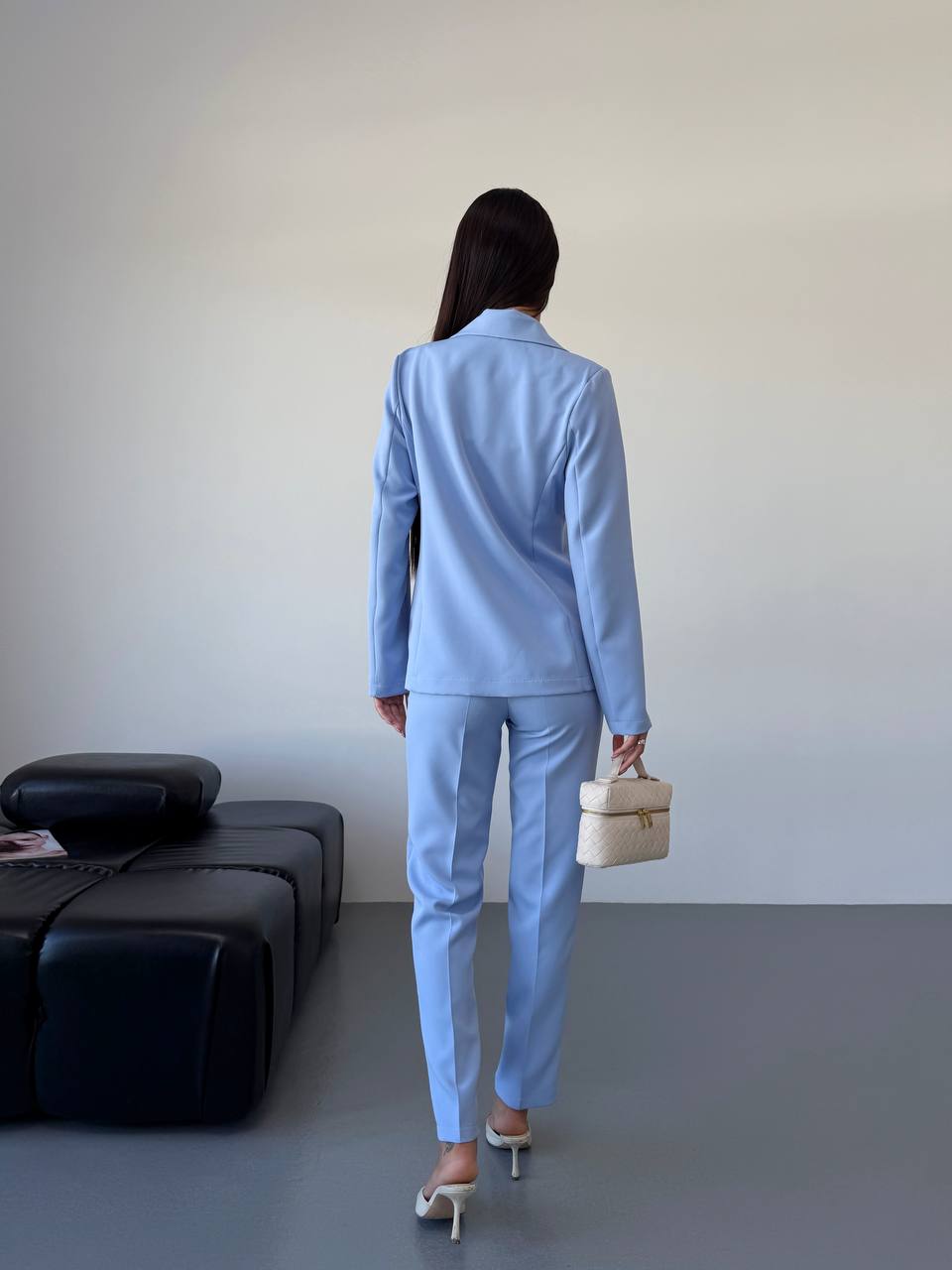 Person wearing a light blue suit standing in a minimalistic room with a black sofa.