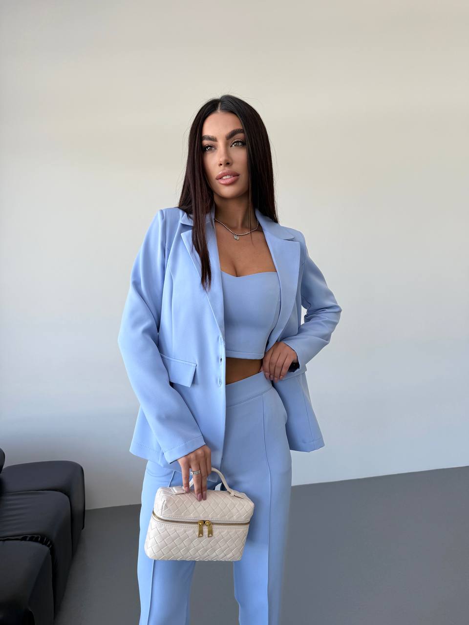 Woman in a light blue suit holding a white handbag against a plain background