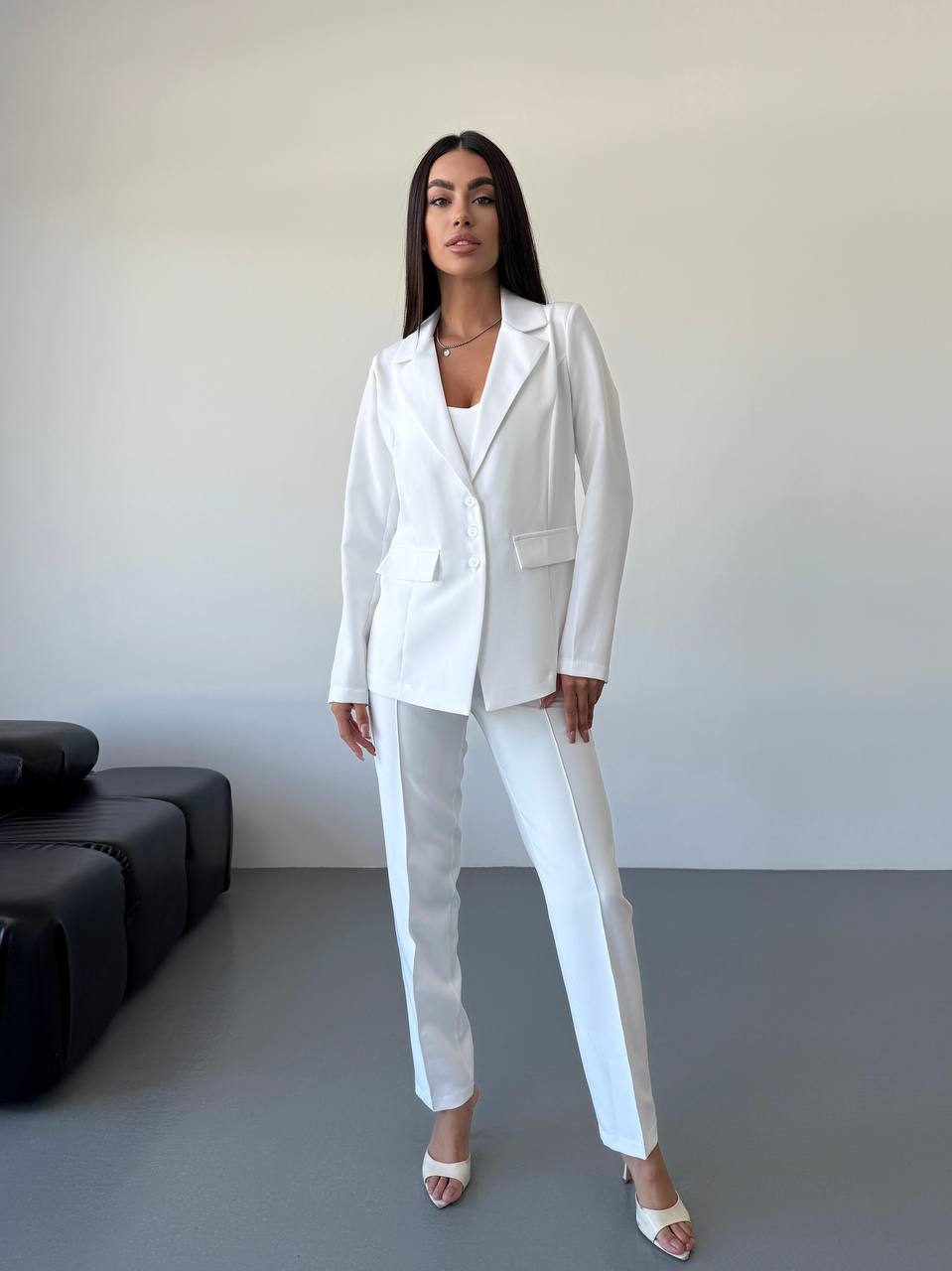 Woman wearing a white suit standing in a minimalistic room with a black sofa.