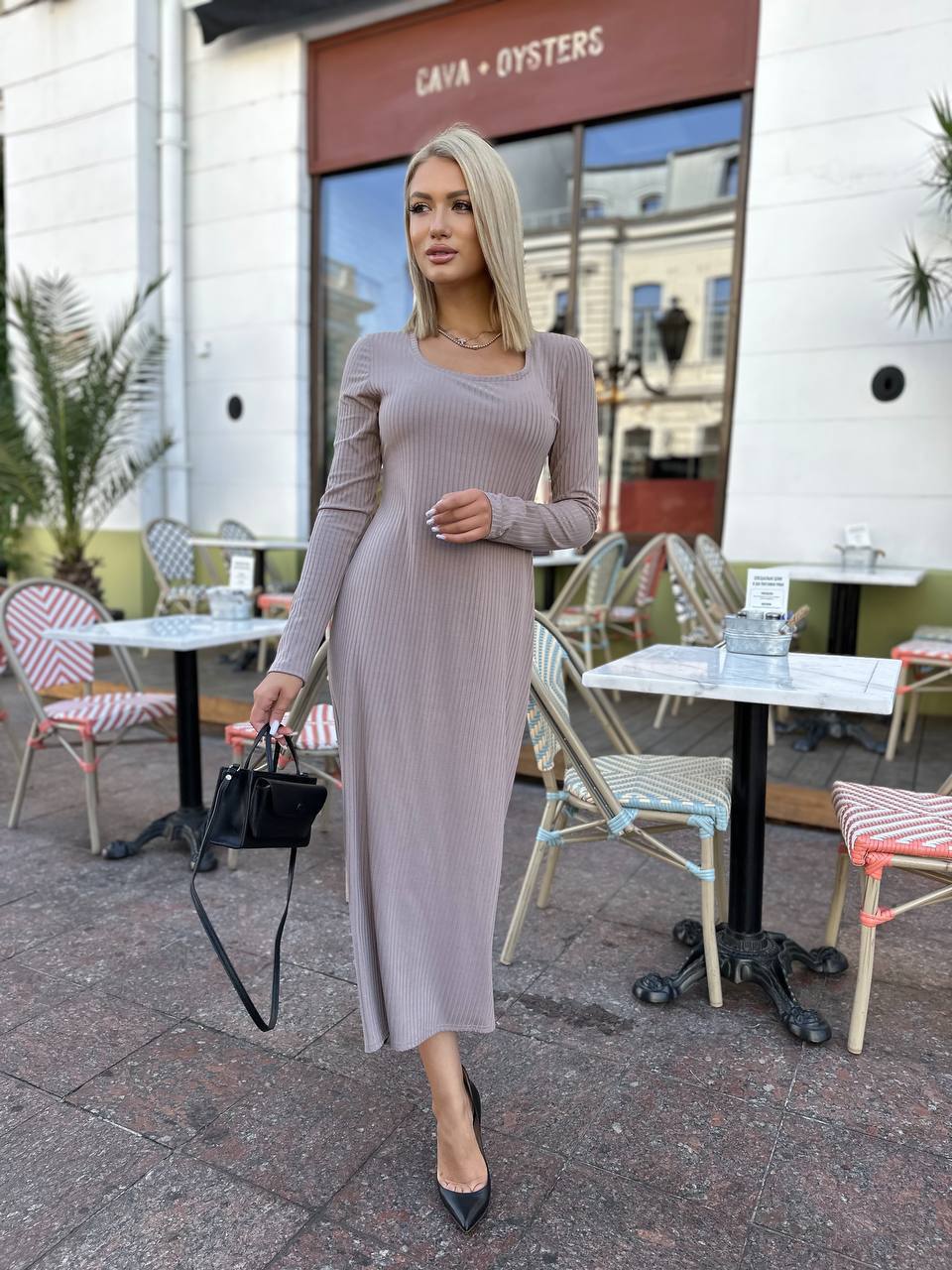 Woman in a long-sleeve dress standing outdoors near a restaurant with tables and chairs.