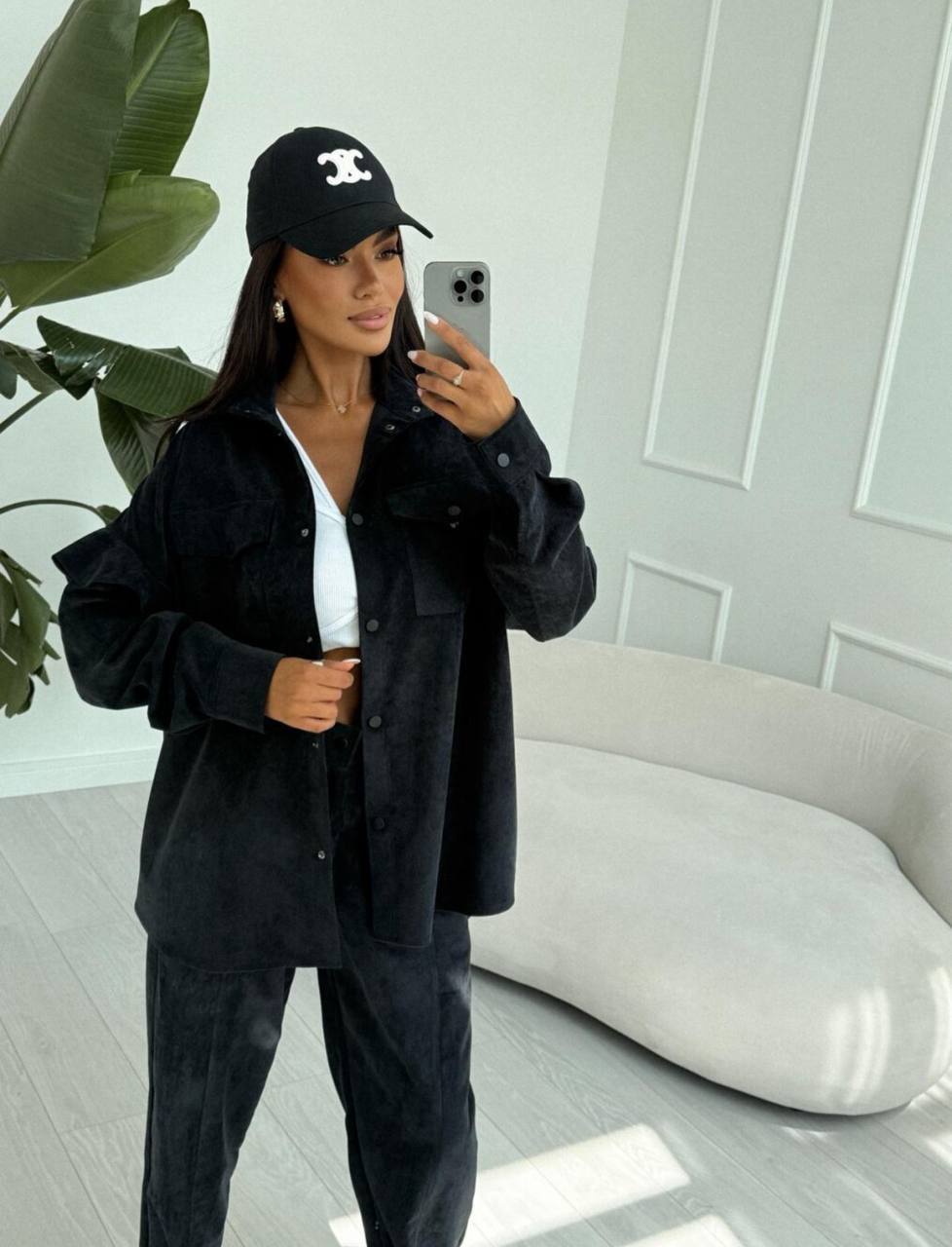 Person taking a mirror selfie wearing a black outfit with a white shirt in a minimalistic room.