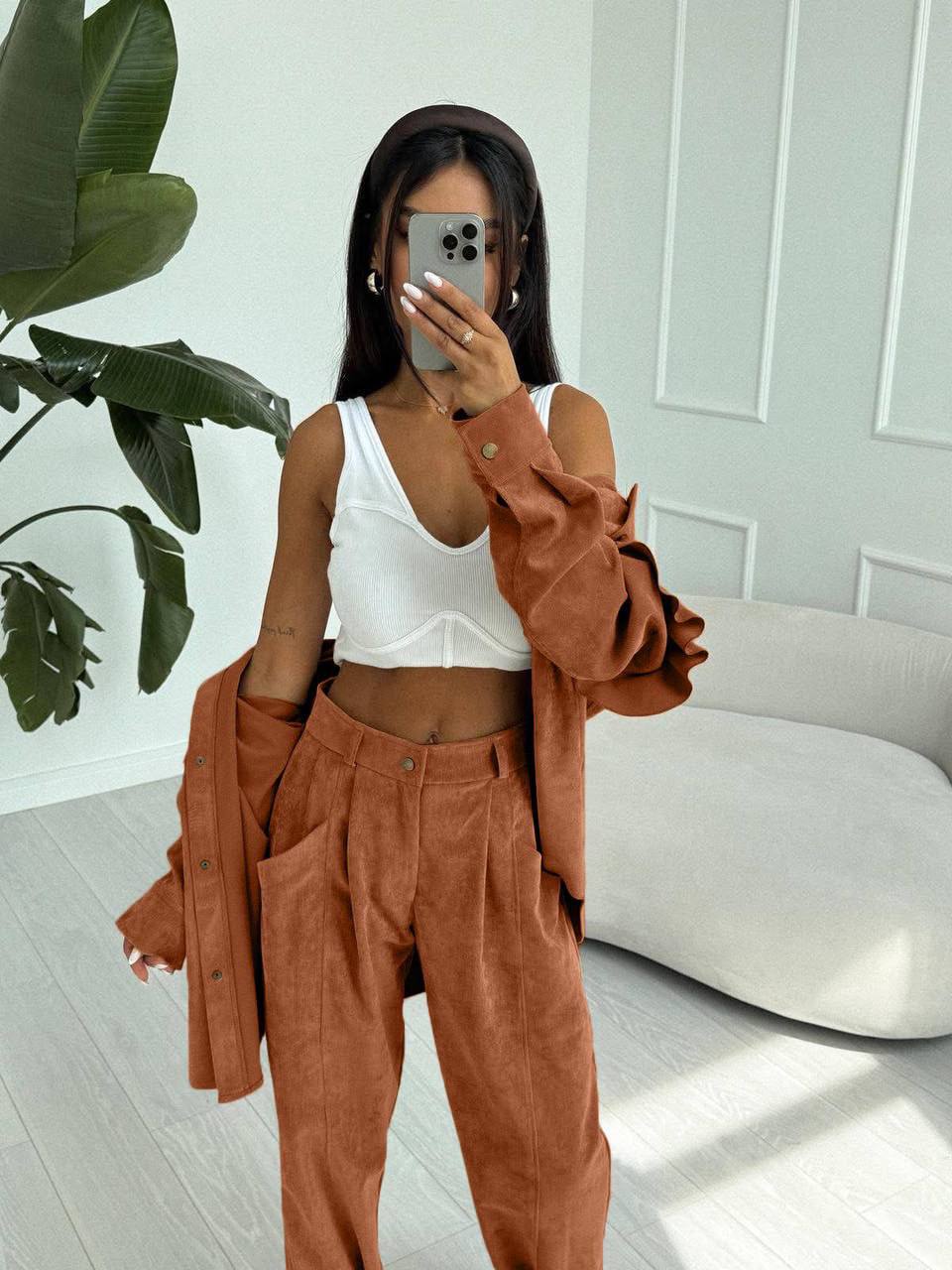 Person wearing a white top and brown pants with a jacket in a room with a plant and white wall.