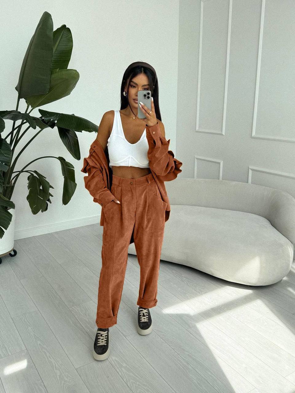 Woman taking a mirror selfie wearing a white top and brown pants in a modern room.