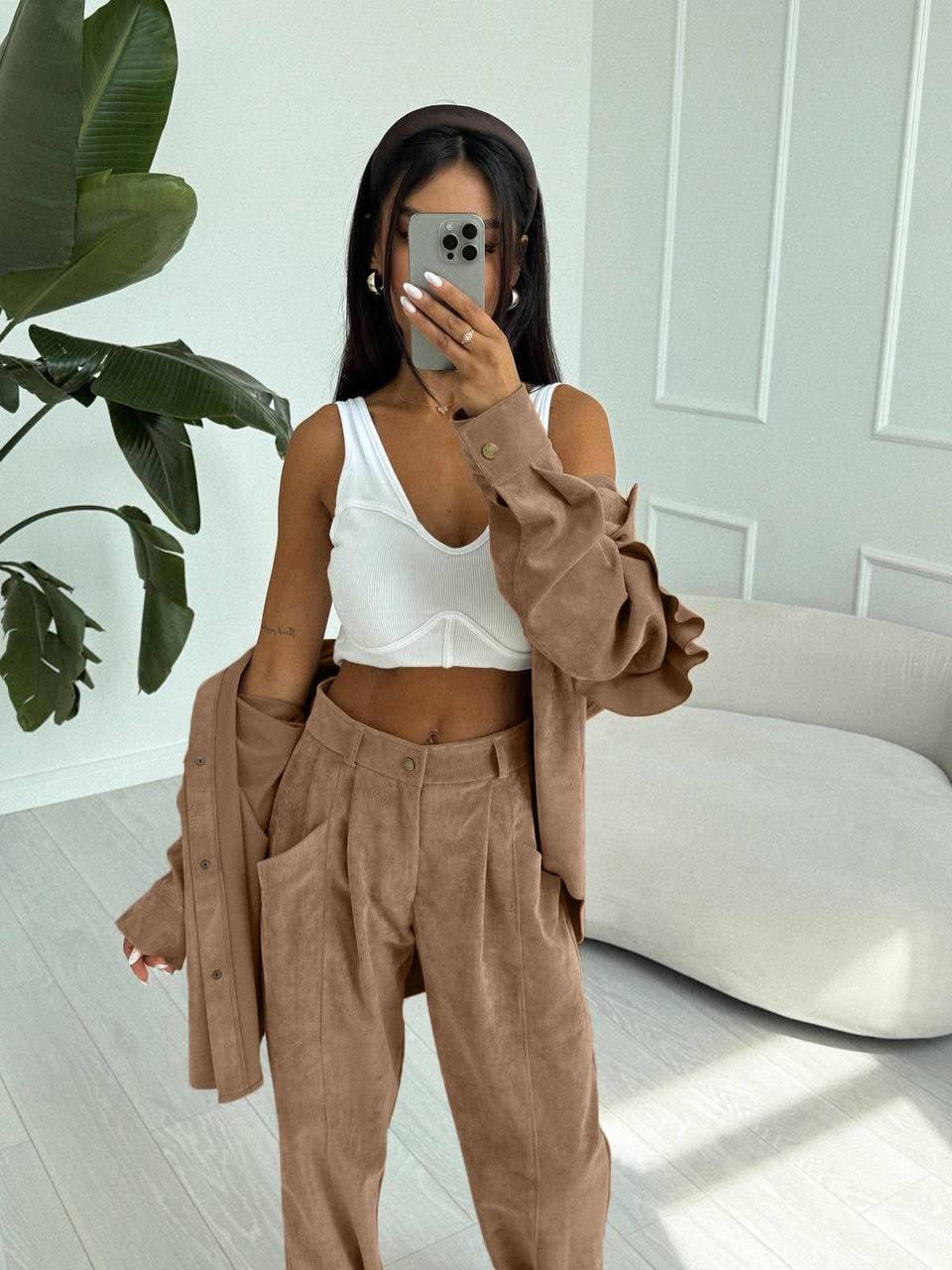 Person wearing a white top and brown pants with a beige cardigan in a room with a plant and sofa.