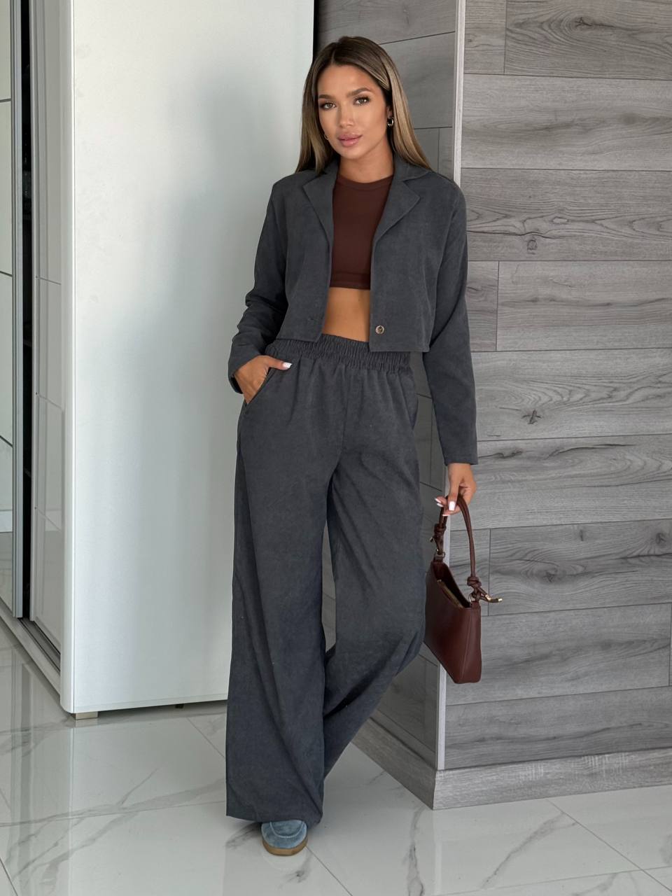 Woman in gray outfit holding a brown handbag against a modern interior background