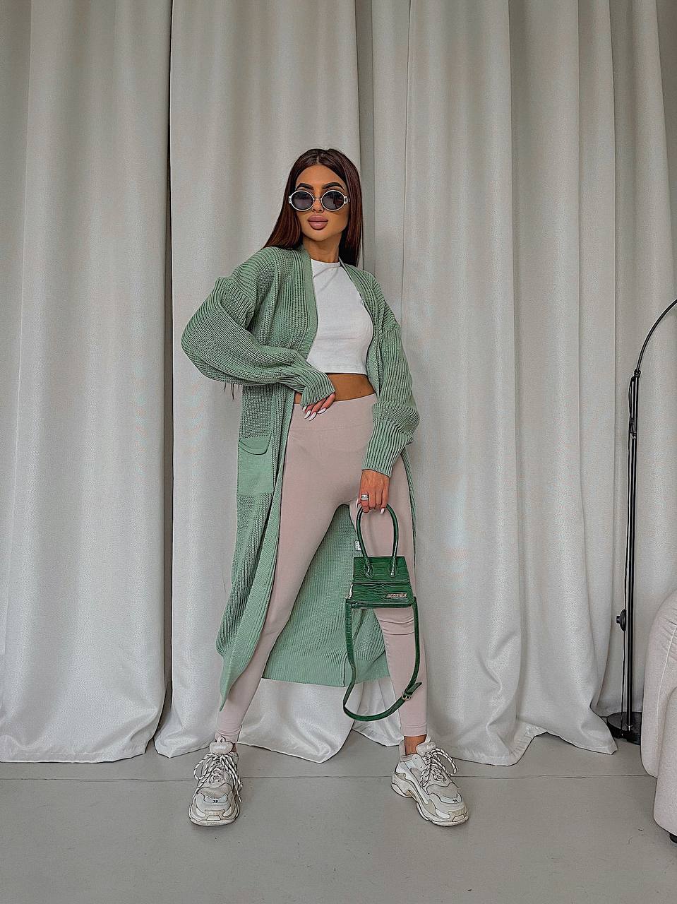 Woman in green cardigan and white pants holding a green handbag against a white curtain background