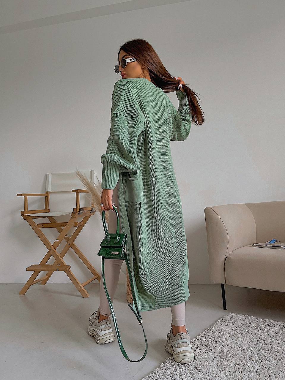 Woman in a long green cardigan holding a green handbag in a room with a chair and sofa.