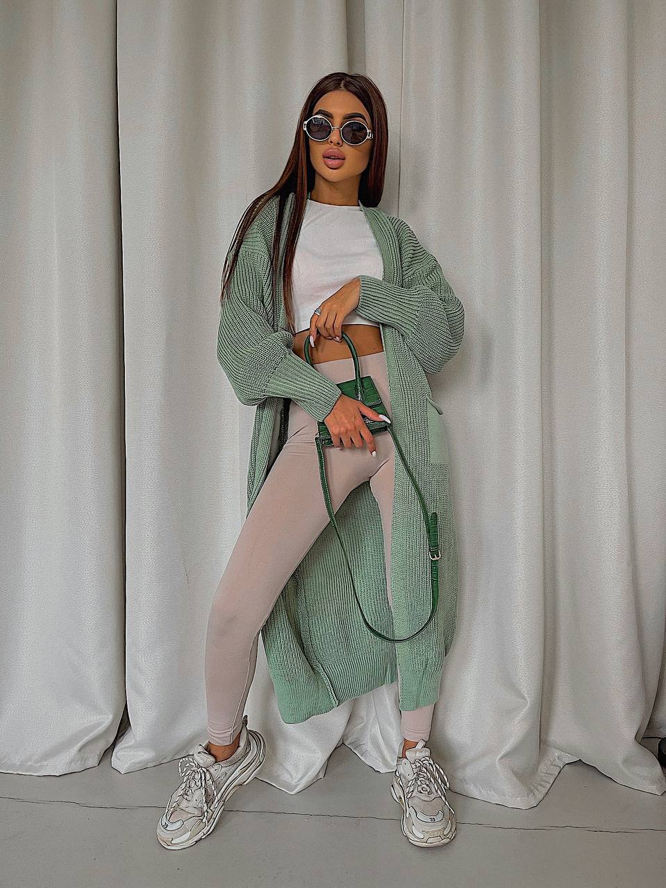 Woman wearing a green cardigan, white top, and leggings against a plain background
