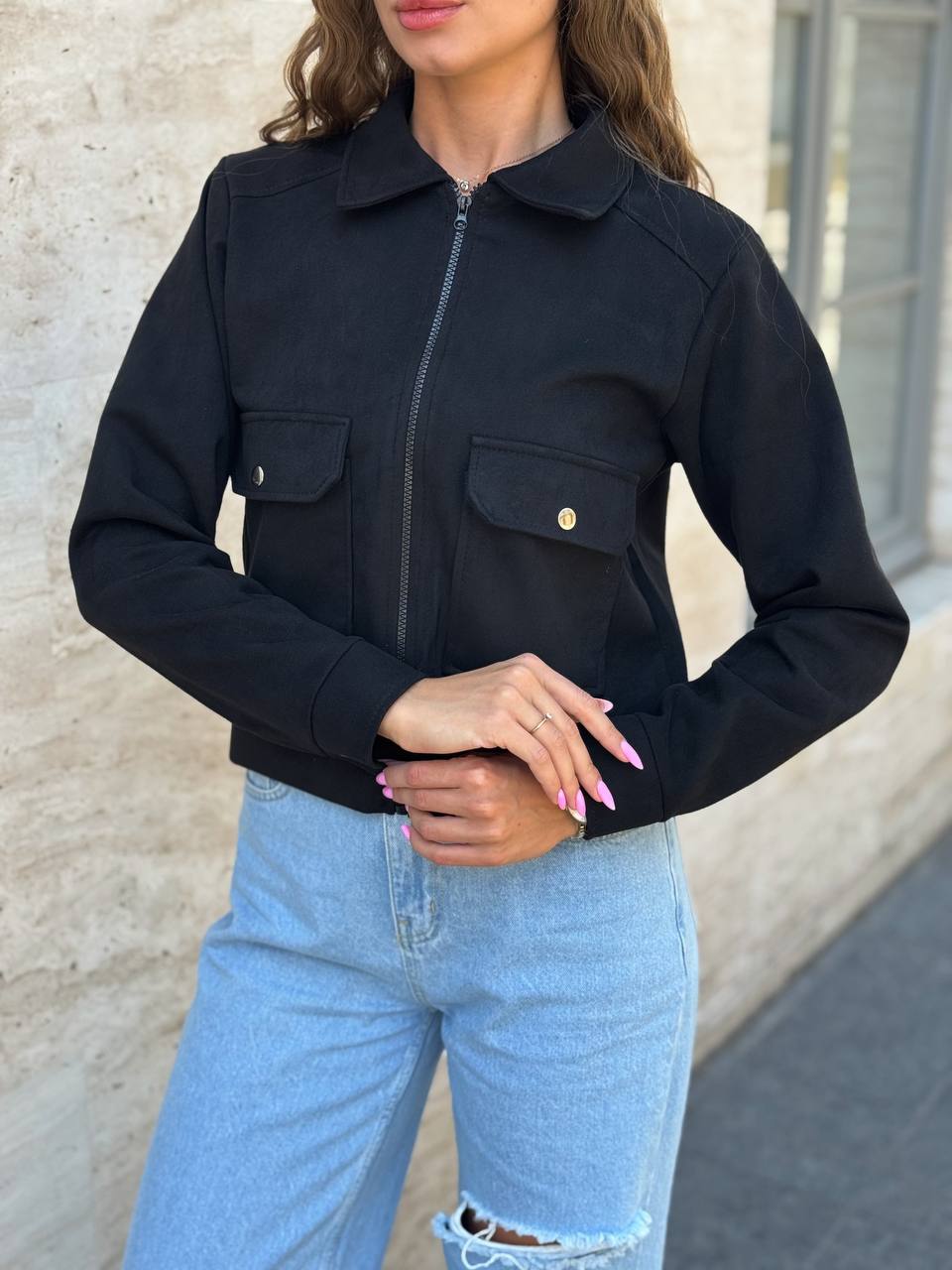 Person wearing a black jacket and light blue jeans against a neutral wall.