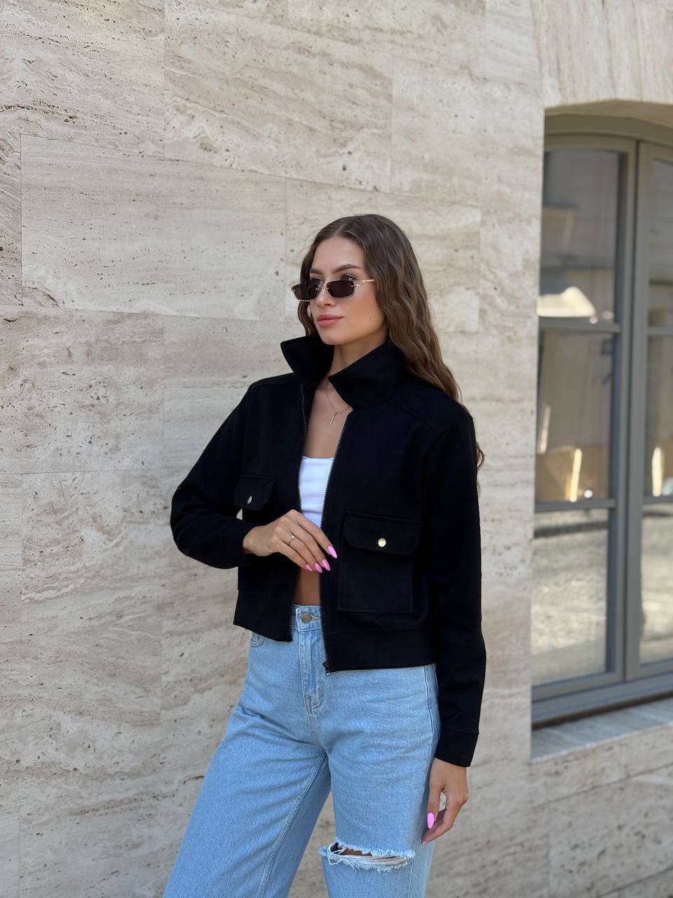 Woman wearing a black jacket and light blue jeans standing against a stone wall.