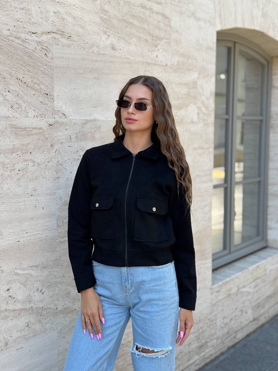 Woman wearing a black jacket and light blue jeans standing against a beige wall.