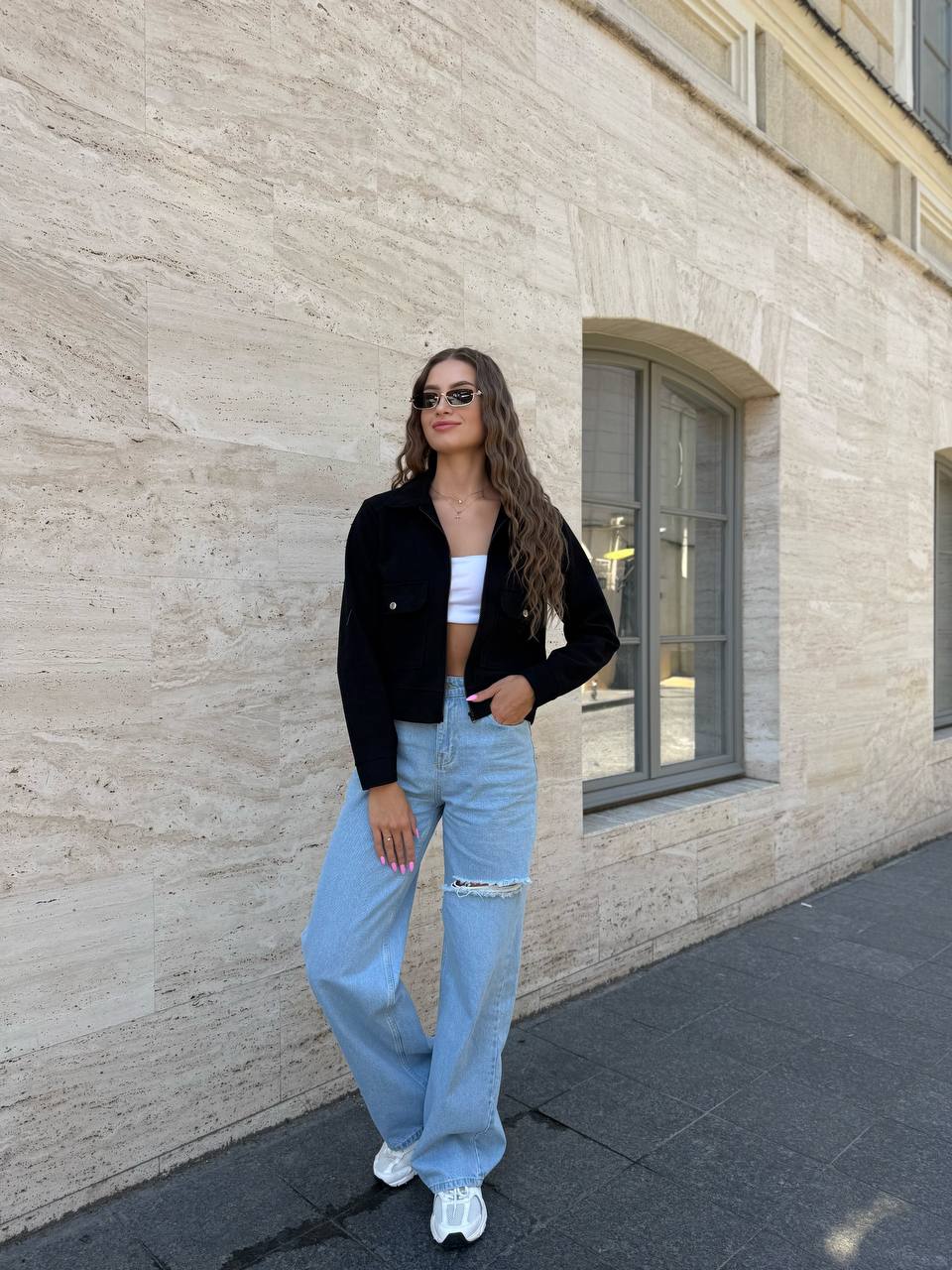 Woman in black jacket, white top, and light blue jeans standing against a stone wall.