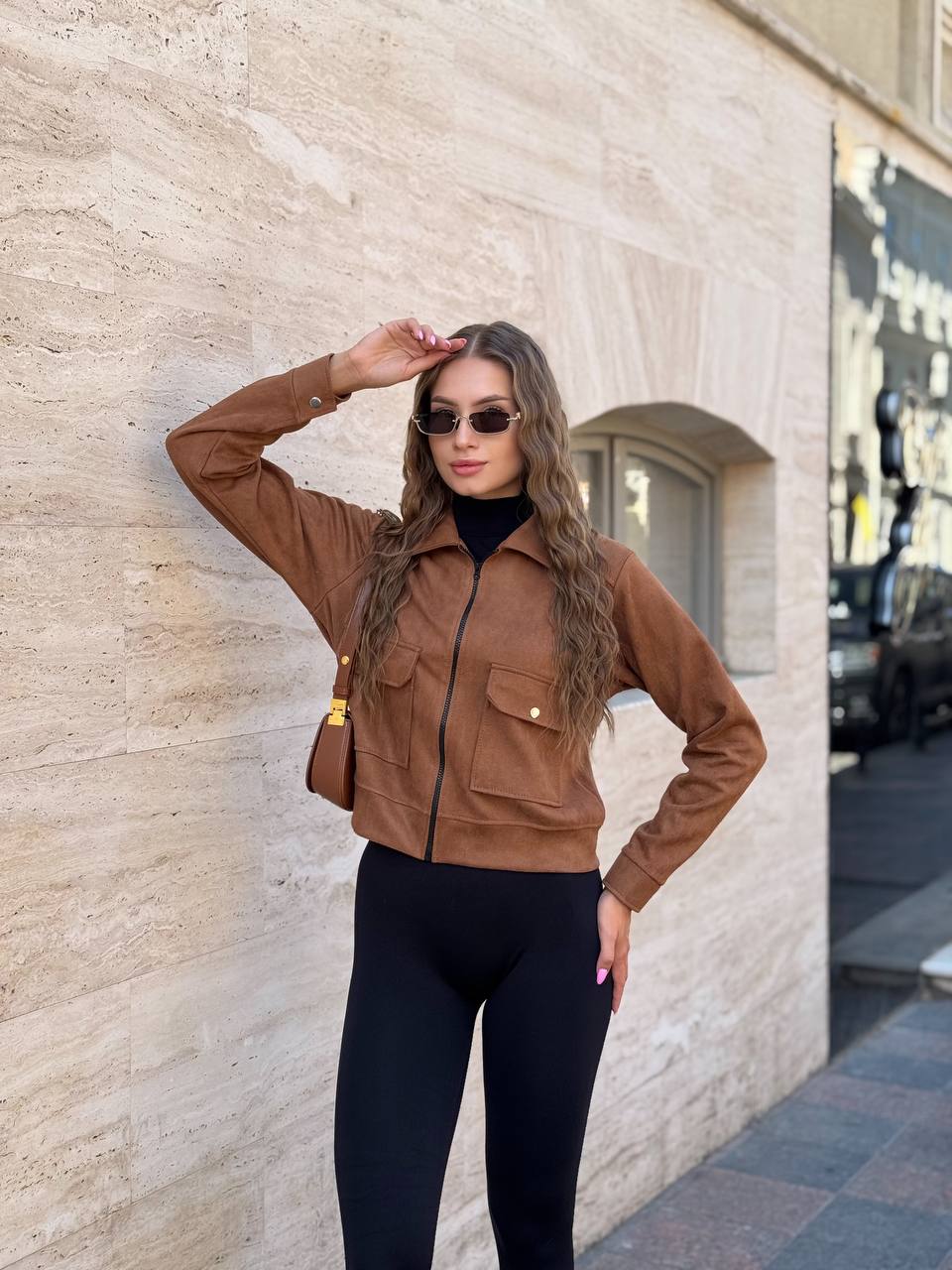 Woman in a brown jacket and black leggings standing against a stone wall.