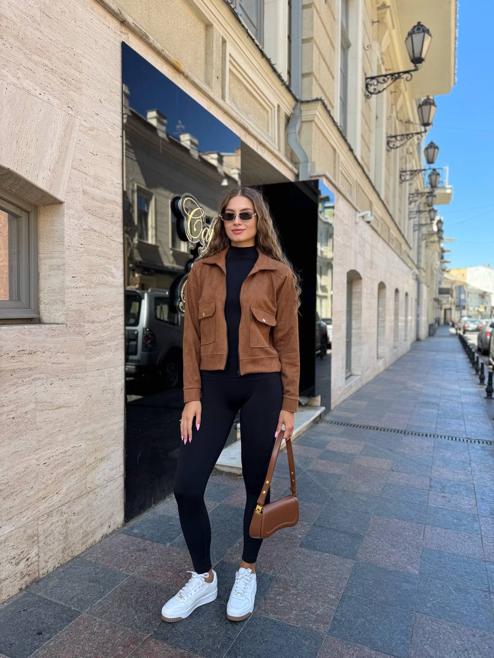Woman in a brown jacket and black leggings standing on a city street.