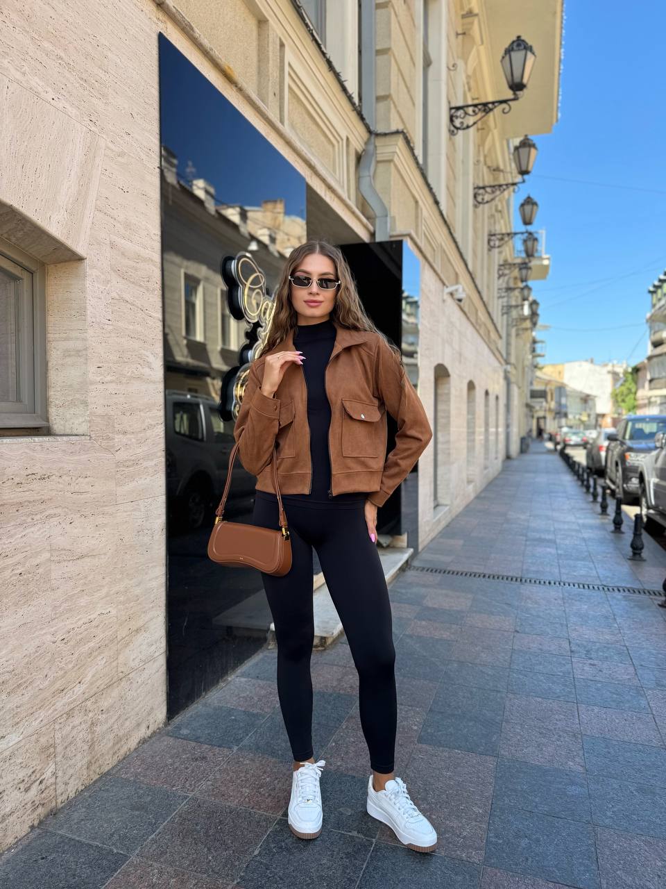 Woman in a brown jacket and black outfit standing on a city street.