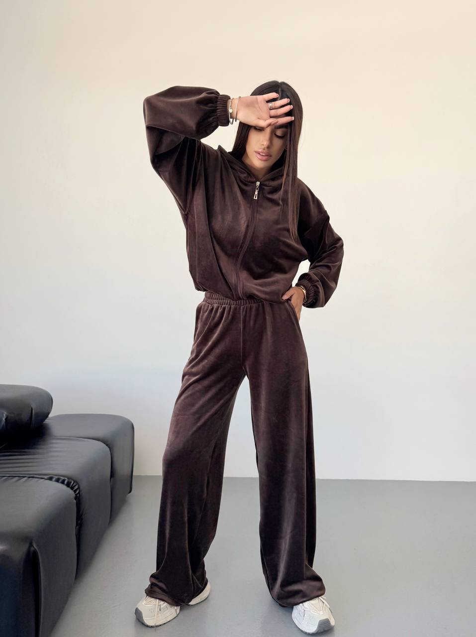 Person wearing a brown tracksuit standing in a minimalistic room with a gray sofa.