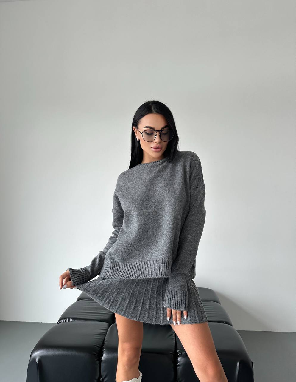 Woman wearing a gray sweater and skirt sitting on a black leather couch against a plain background