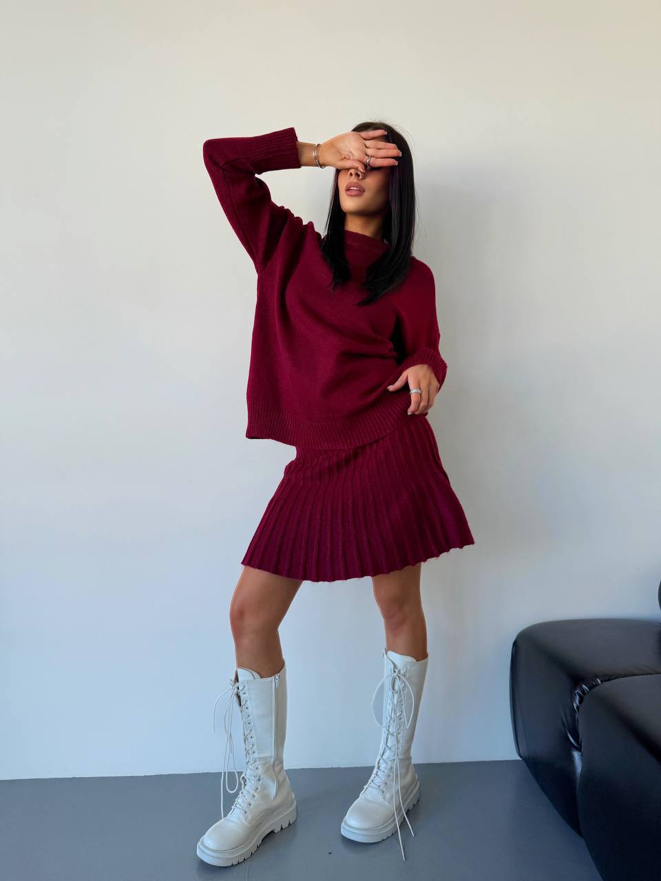 Person wearing a maroon sweater and skirt with white boots against a plain background