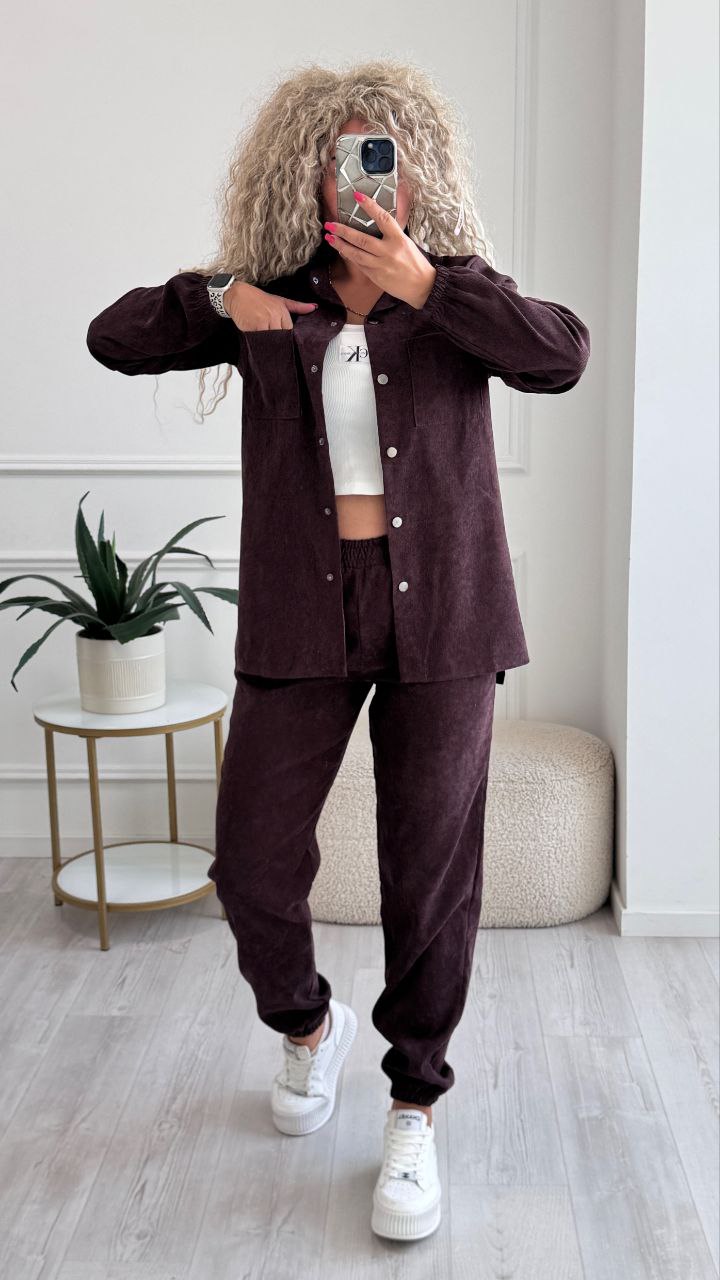 Person wearing a brown outfit with a white top, standing in a room with a plant and small table.