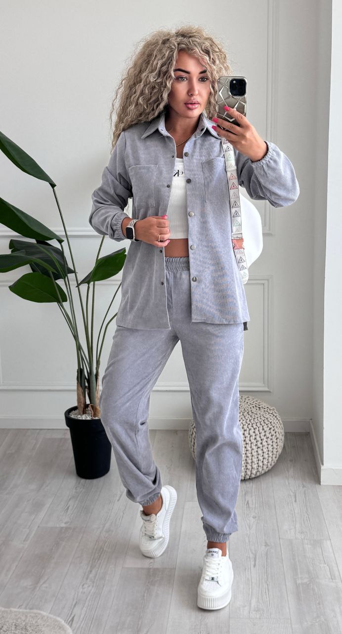 Woman taking a mirror selfie wearing a gray tracksuit in a room with a plant and rug.