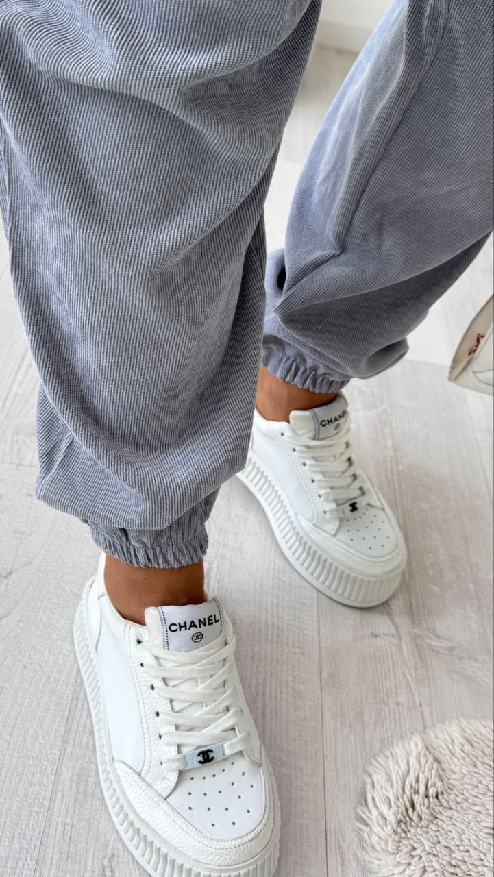 White sneakers with a brand logo worn with gray pants on a light wooden floor.