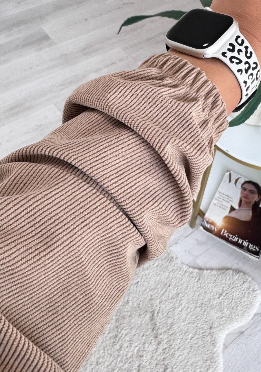 Person wearing a beige ribbed sweater with a leopard print smartwatch on a light wooden floor.
